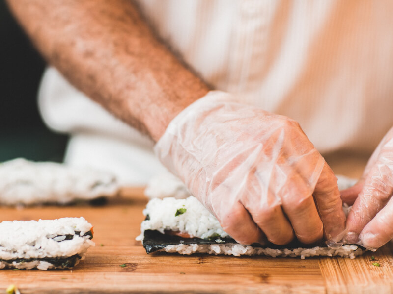 A person wearing plastic gloves rolling sushi on a wooden chopping board. 