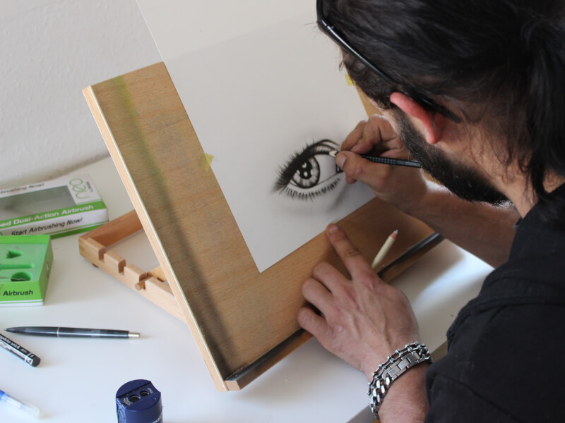 A person is sat at a drawing board, drawing a picture of an eye