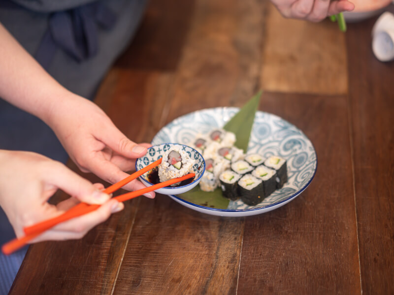A close up of a person holding sushi with chopsticks, dipping it into a small bowl of sauce