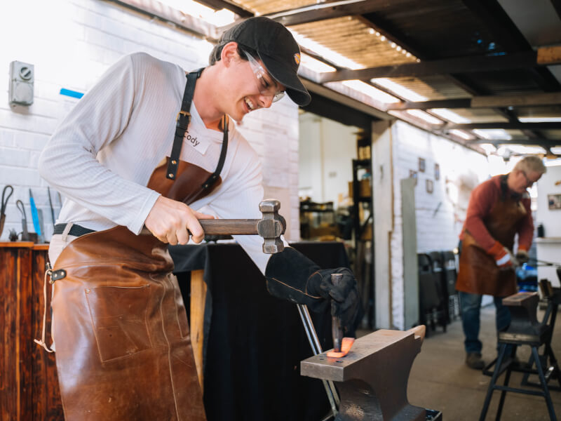 A man is wearing an apron and hammering a piece of molten metal into shape