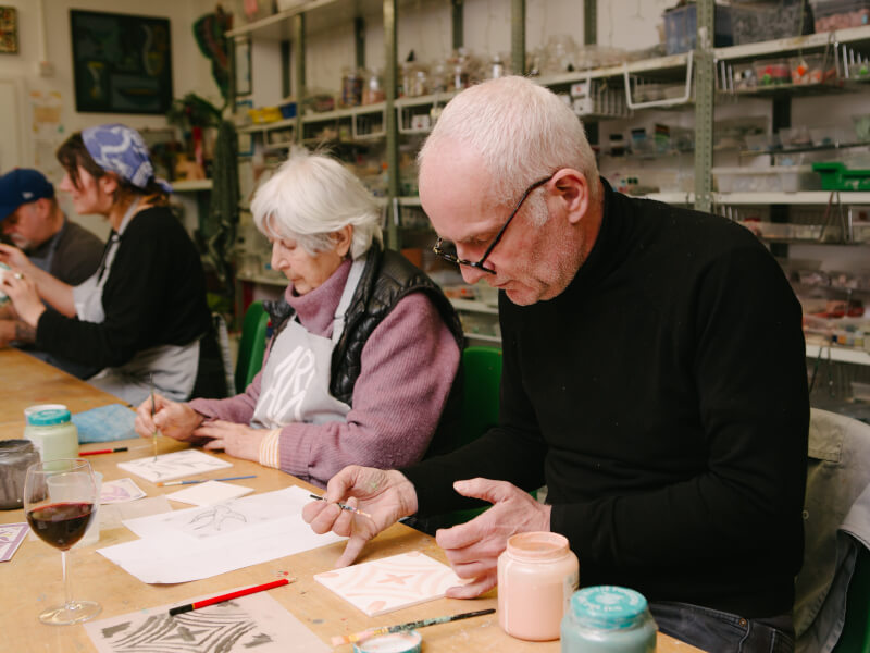 A man and woman sit at a table, painting designs on white tiles