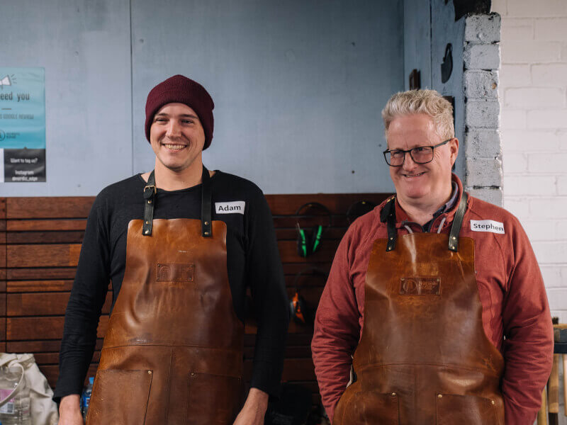An older and younger man are standing in a workshop, wearing aprons and preparing to learn craft skills