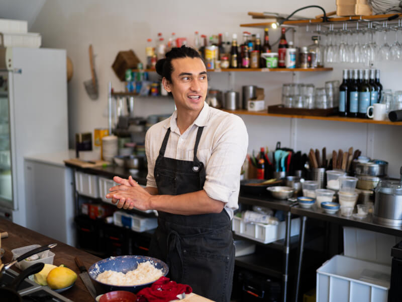 A man is standing in a kitchen wearing an apron, in front of a table with various bowls of ingredients on it
