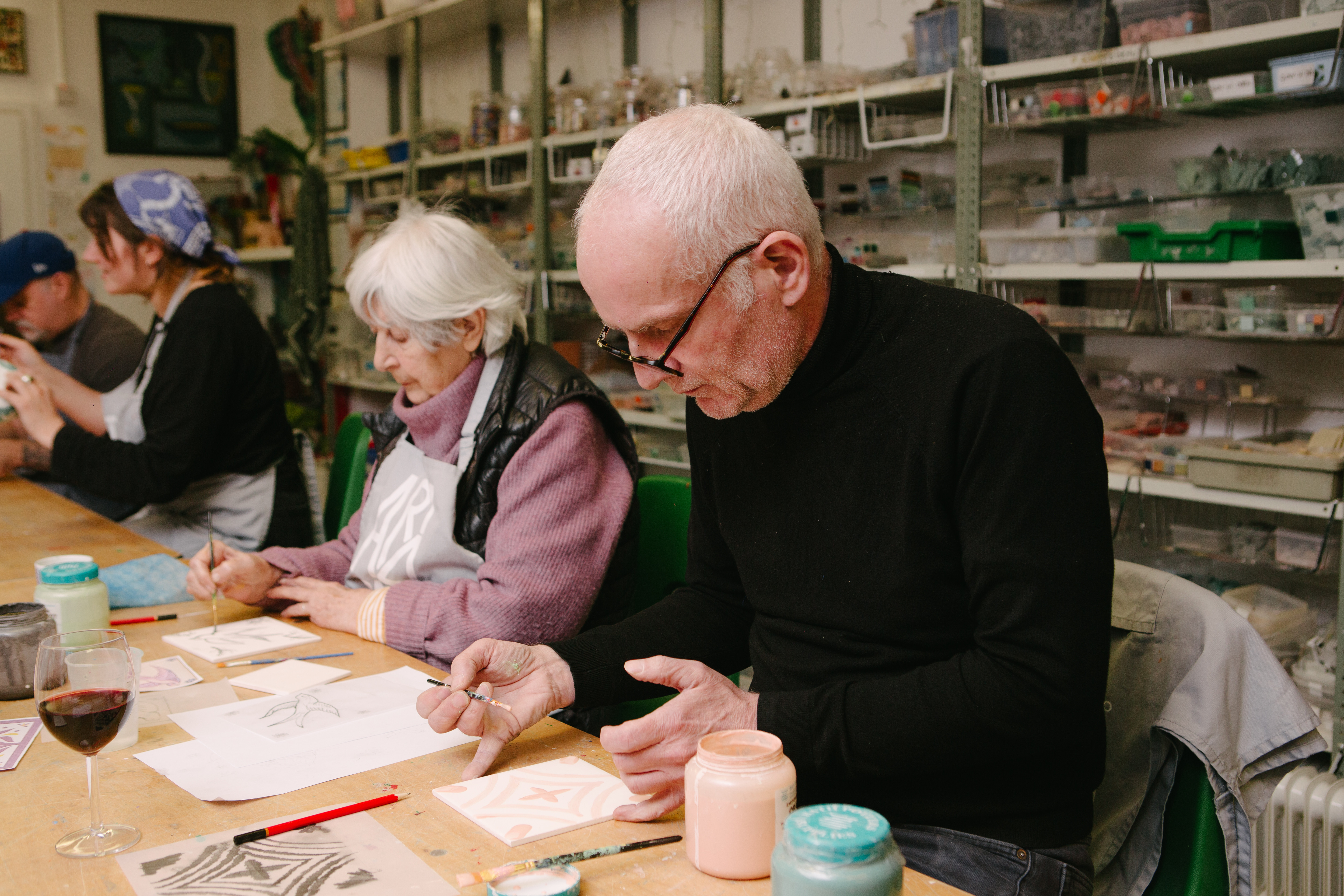 A man and woman are sitting at a table, painting patterns on ceramic tiles with pink paint
