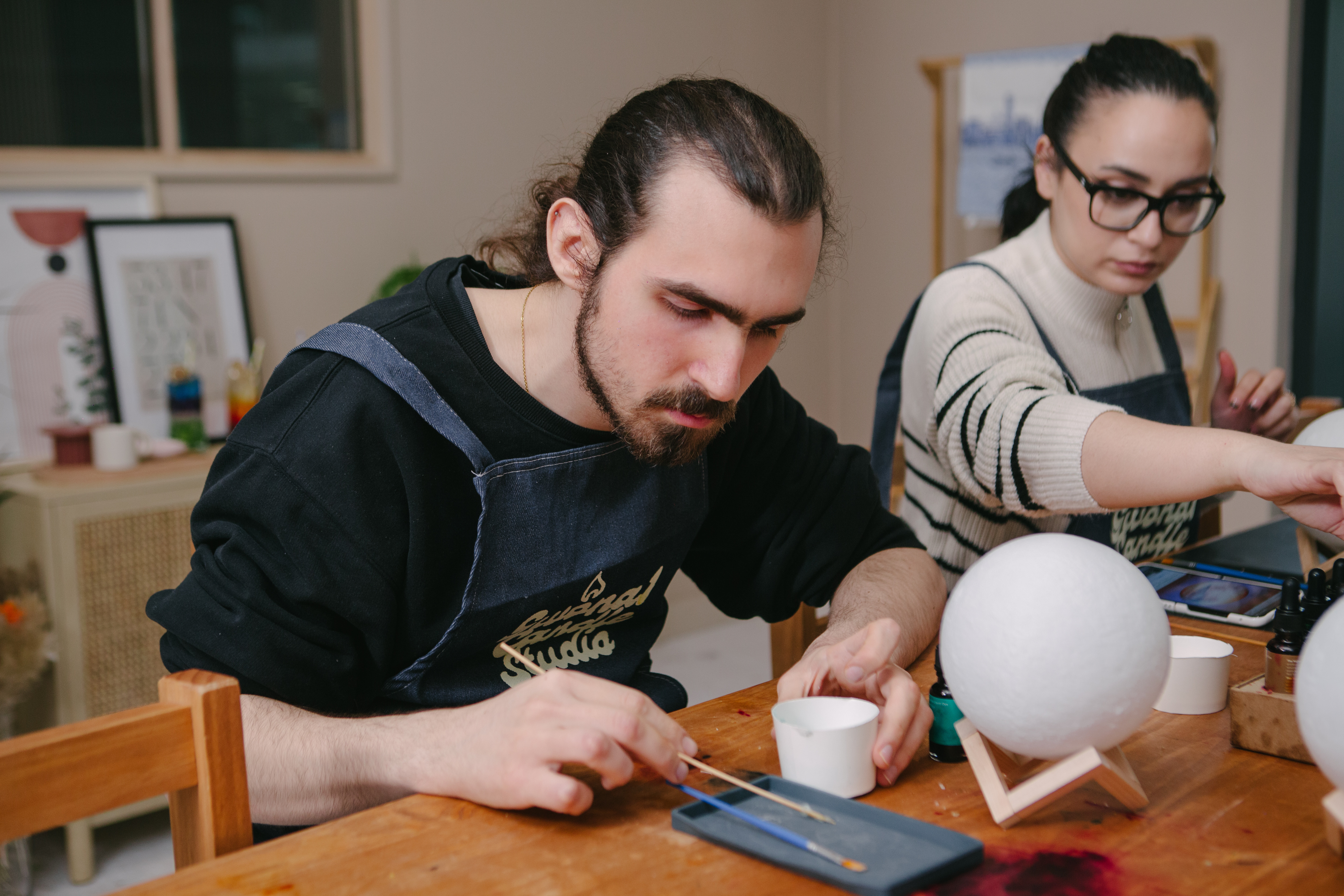 A man and woman are sitting at a table, making a wax globe lamp