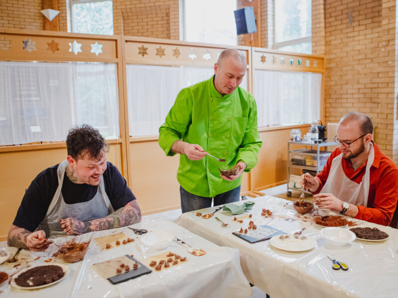 Two men sit at a table at a chocolate making class, being taught by a chef in a green jacket