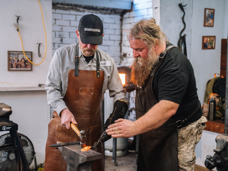 Two men are shaping molten metal with hammers at a metal working class