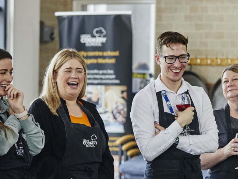 Three people at a cooking class from Leeds Cookery School are laughing and watching a demonstration