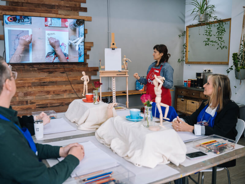 A group of people at a drawing class are sitting around a table, watching a video about drawing