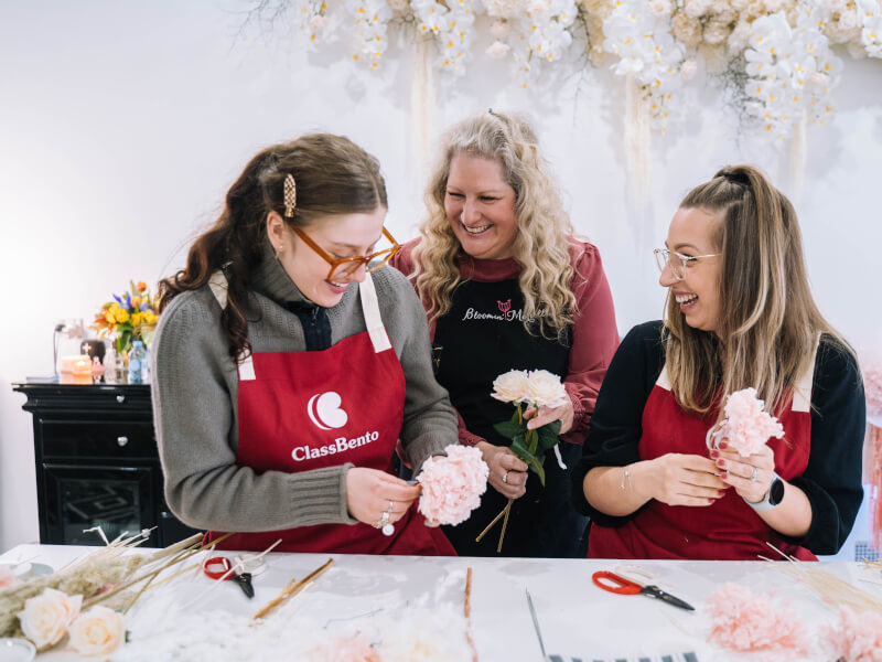 Three women are smiling and holding pale pink flowers
