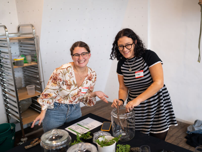 Two women are standing in front of a table filled with plants, soil and containers