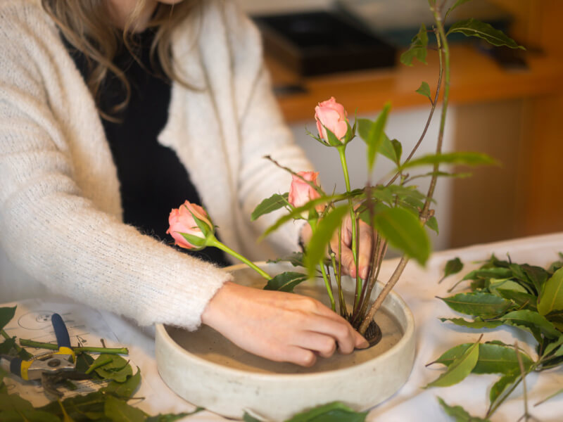 A person is creating an ikebana flower arrangement with pink roses in a beige stoneware dish