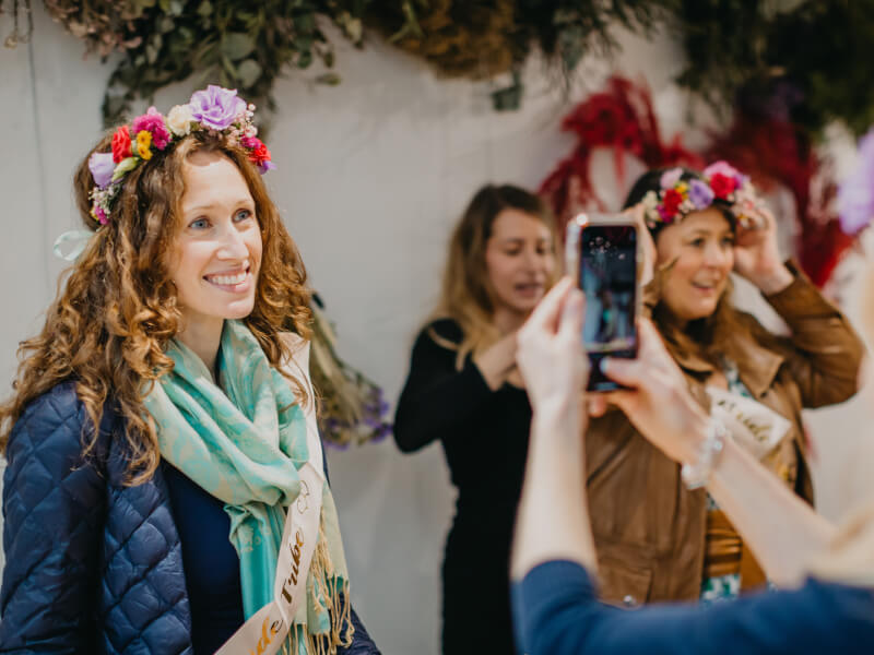 A woman in a flower crown is smiling and having her photo taken by another woman