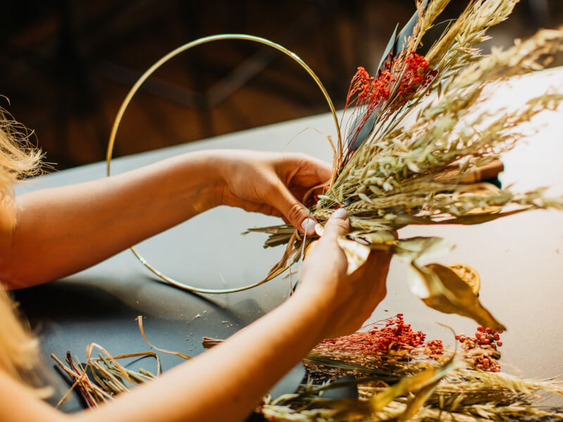 A close up of a person making a dried flower wreath from red flowers and neutral foliage