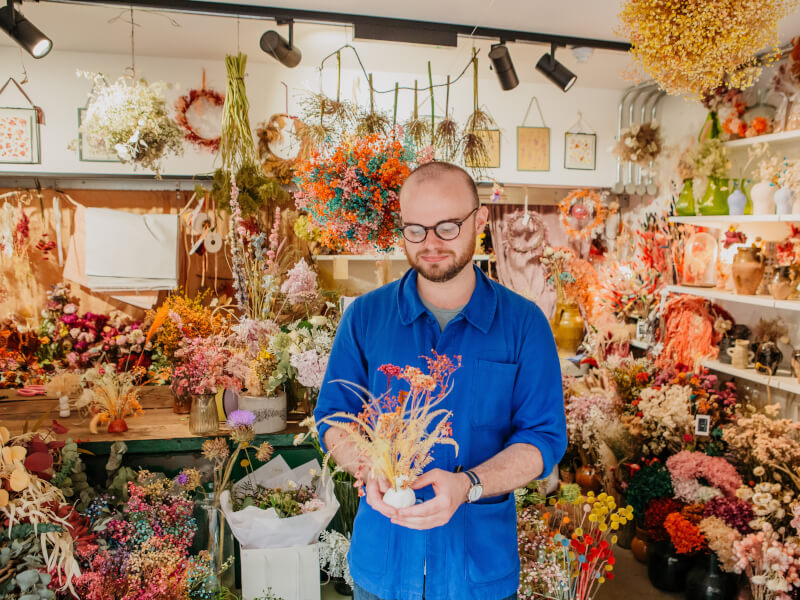 A man in a blue shirt is standing in a room full of flowers, holding an ikebana floral arrangement