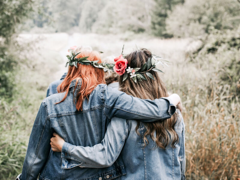 Two women photographed from behind are wearing flower crowns and walking through a field
