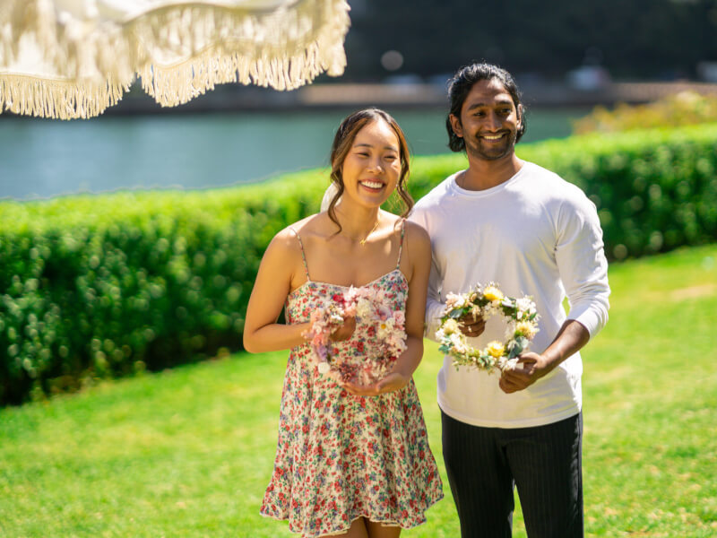 A man and woman are standing in a sunny outdoor space, holding handmade flower wreaths