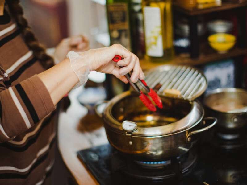 A close up of a person holding red tongs and heating oil in a pan ready to deep fry churros
