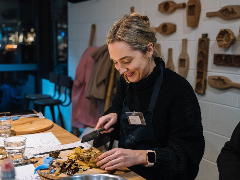 A woman sits at a wooden table, slicing large mushrooms on a chopping board