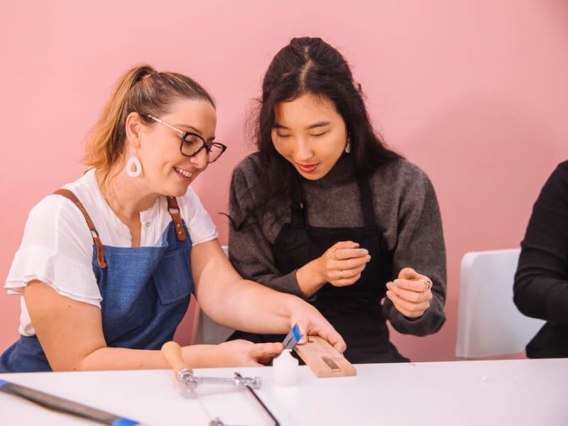 Two women against a pink wall smile at a craft table