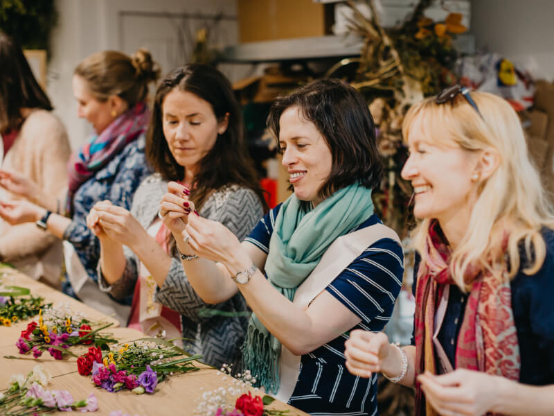 A group of women are sitting round a big table, learning how to make flower crowns