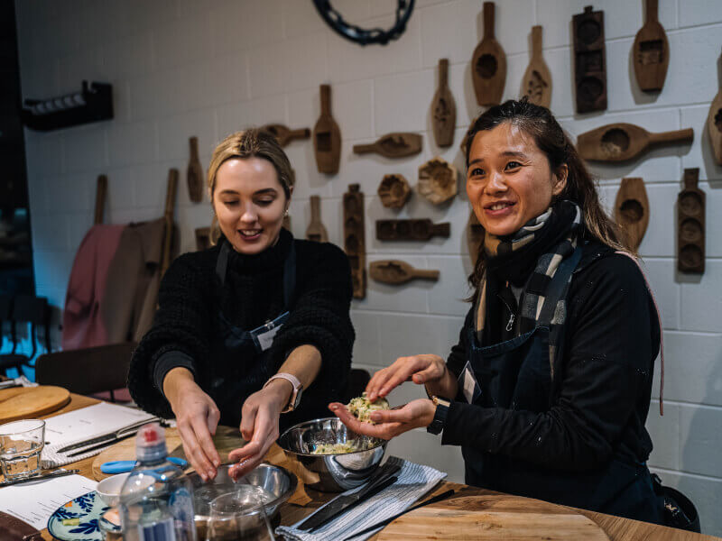 Two women in a cooking class are mixing ingredients in a bowl with their hands and smiling