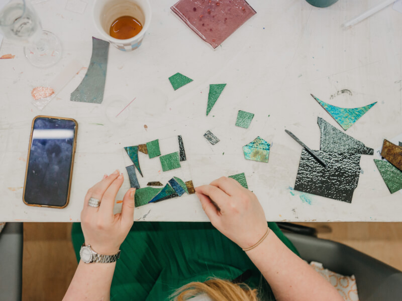 A top down shot of a person arranging blue and green glass pieces to make a piece of mosaic art 