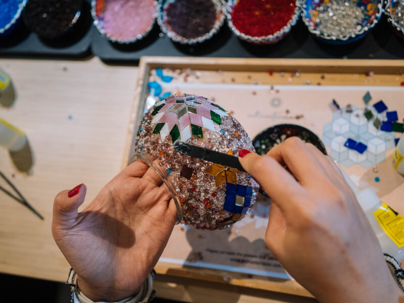 A top down shot of someone sticking brown beads to a colourful mosaic design on a glass jar