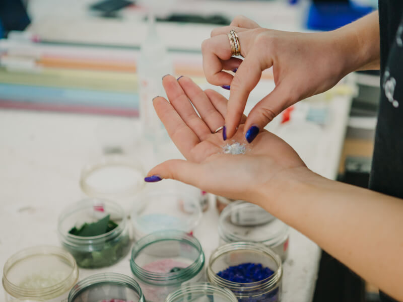 A person is holding small pieces of blue and white glass in their hand and there are pots of coloured glass pieces on the table in front of them