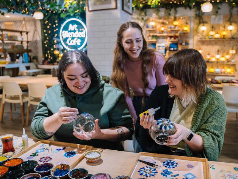 Two women are sitting at a table at a mosaic workshop, starting their project while the teacher looks on
