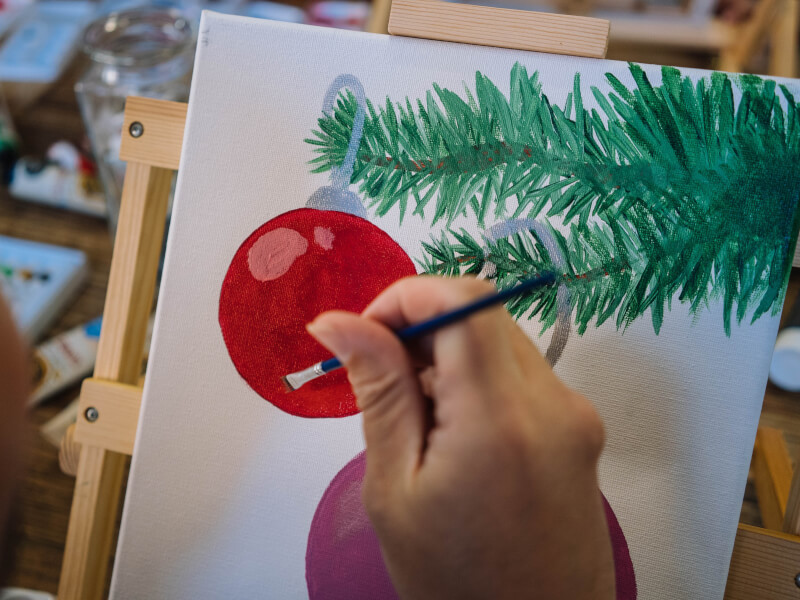 A close up of a person painting a picture of a bauble on a Christmas tree branch
