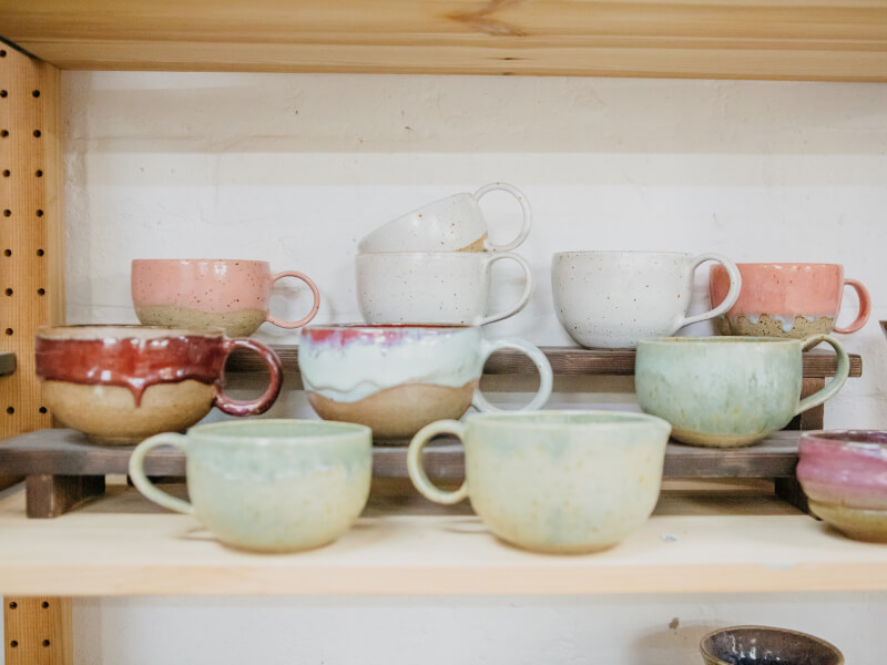 A selection of handmade ceramic cups in different coloured glazes on a wooden shelf