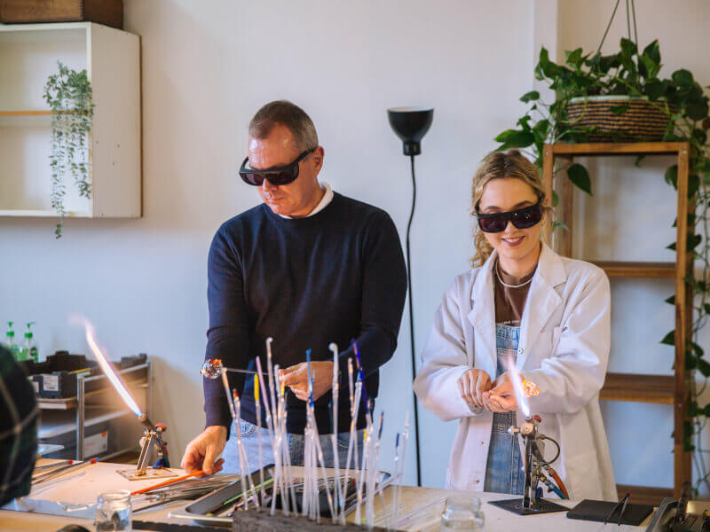 A man and woman are heating and shaping liquid glass using flames