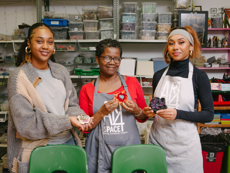 Three women are standing in an art studio holding handmade fabric flowers