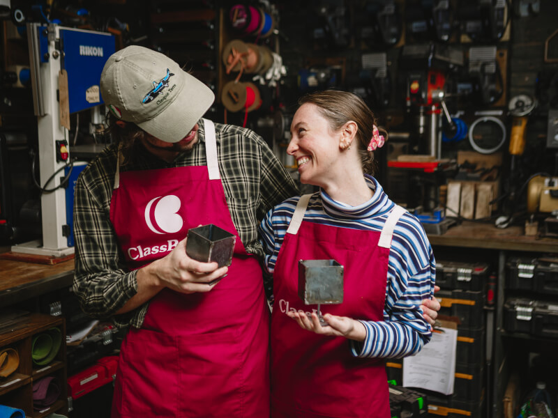 A man and woman are smiling and holding metal boxes they've made at a metalwork class