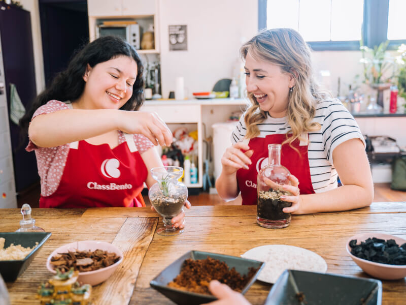 Two women are sitting at a table, adding plants to glass terrariums
