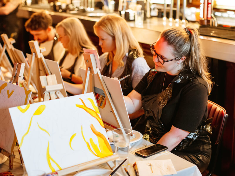 A group of women are sitting at easels on a table at a hen party paint and sip 