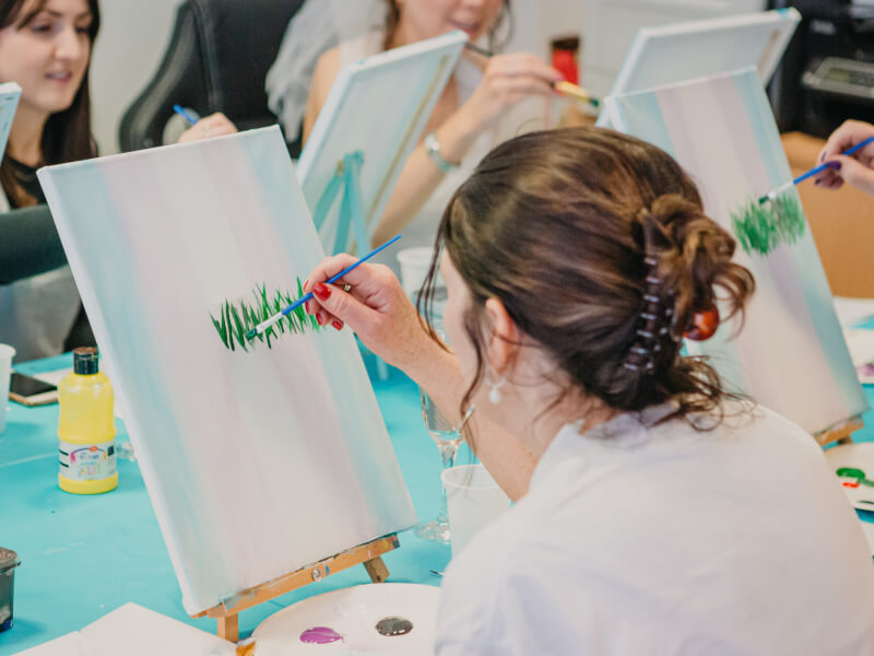 A woman is sitting at an easel, painting grass onto a white canvas