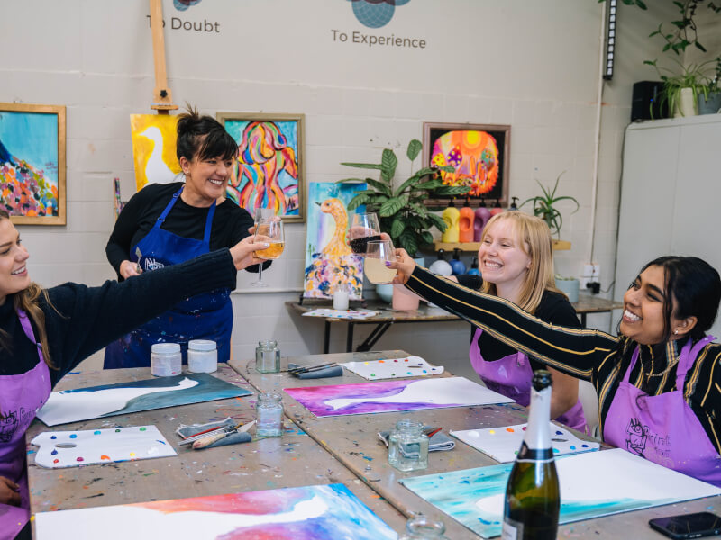 A group of women at a paint and sip class are sitting around a table with canvases, clinking glasses