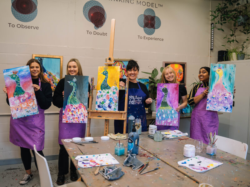 A group of women at a paint and sip hen party are holding up their finished paintings of peacocks