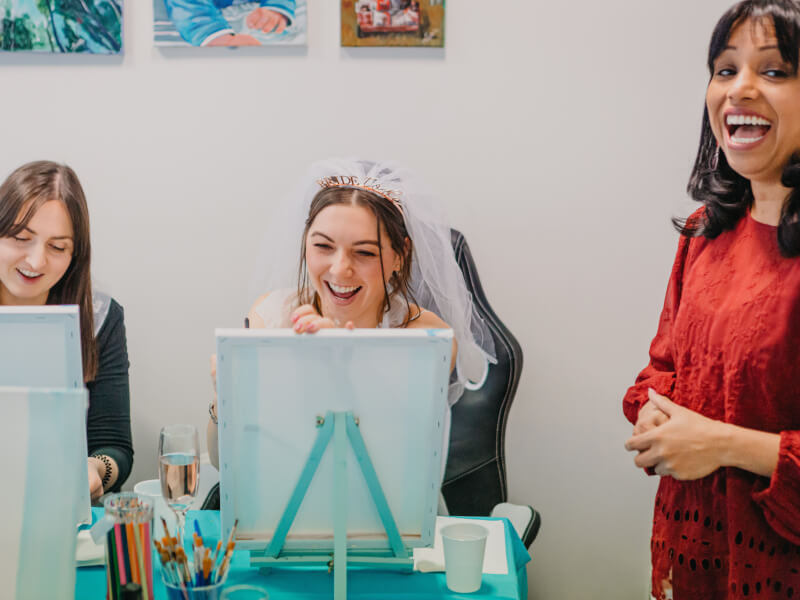 A bride-to-be is sitting next to her friend, wearing a veil, and the art teacher is standing next to them at a paint and sip class