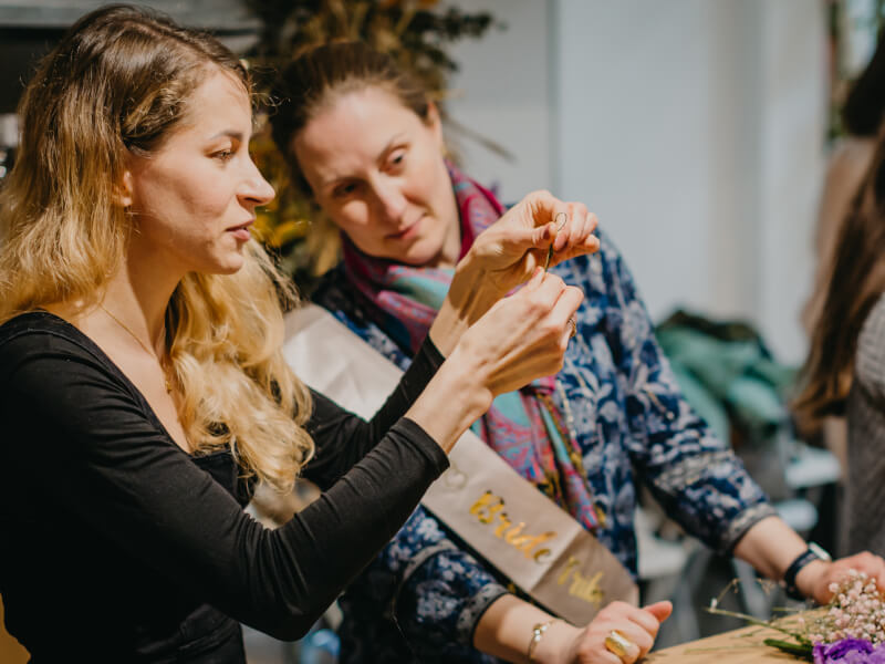 A woman is threading a needle while another woman looks on at a floral crown class