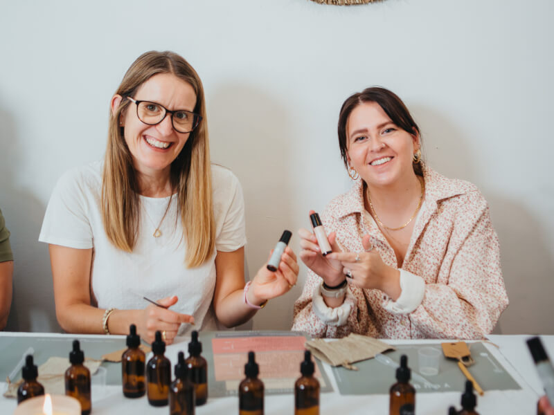 Two smiling women hold up aromatherapy rollerballs at a perfume workshop