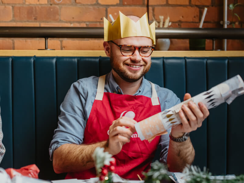 A smiling man is wearing a yellow paper crown and making a DIY Christmas cracker