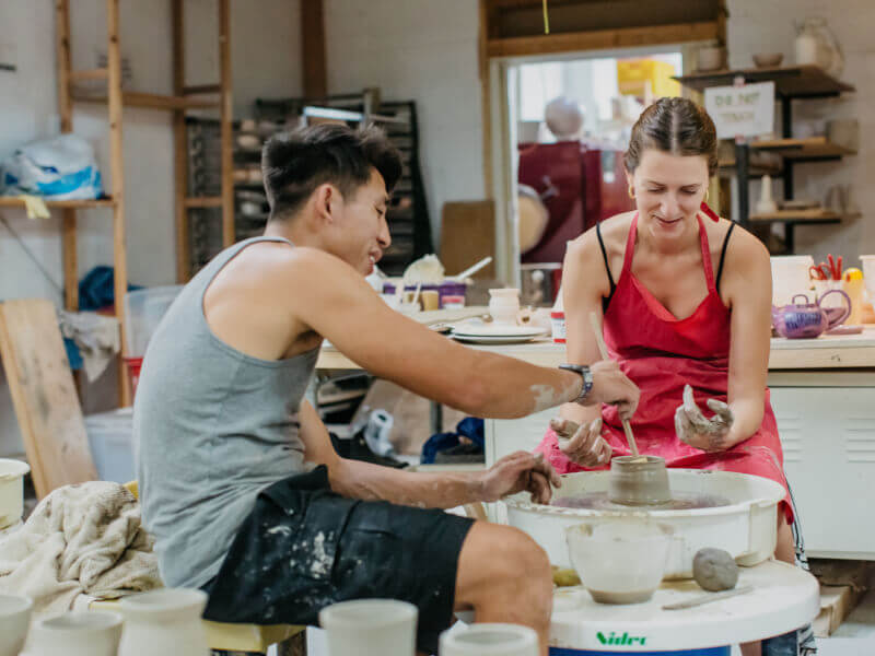 Two people are making a small pot from clay on a pottery wheel 