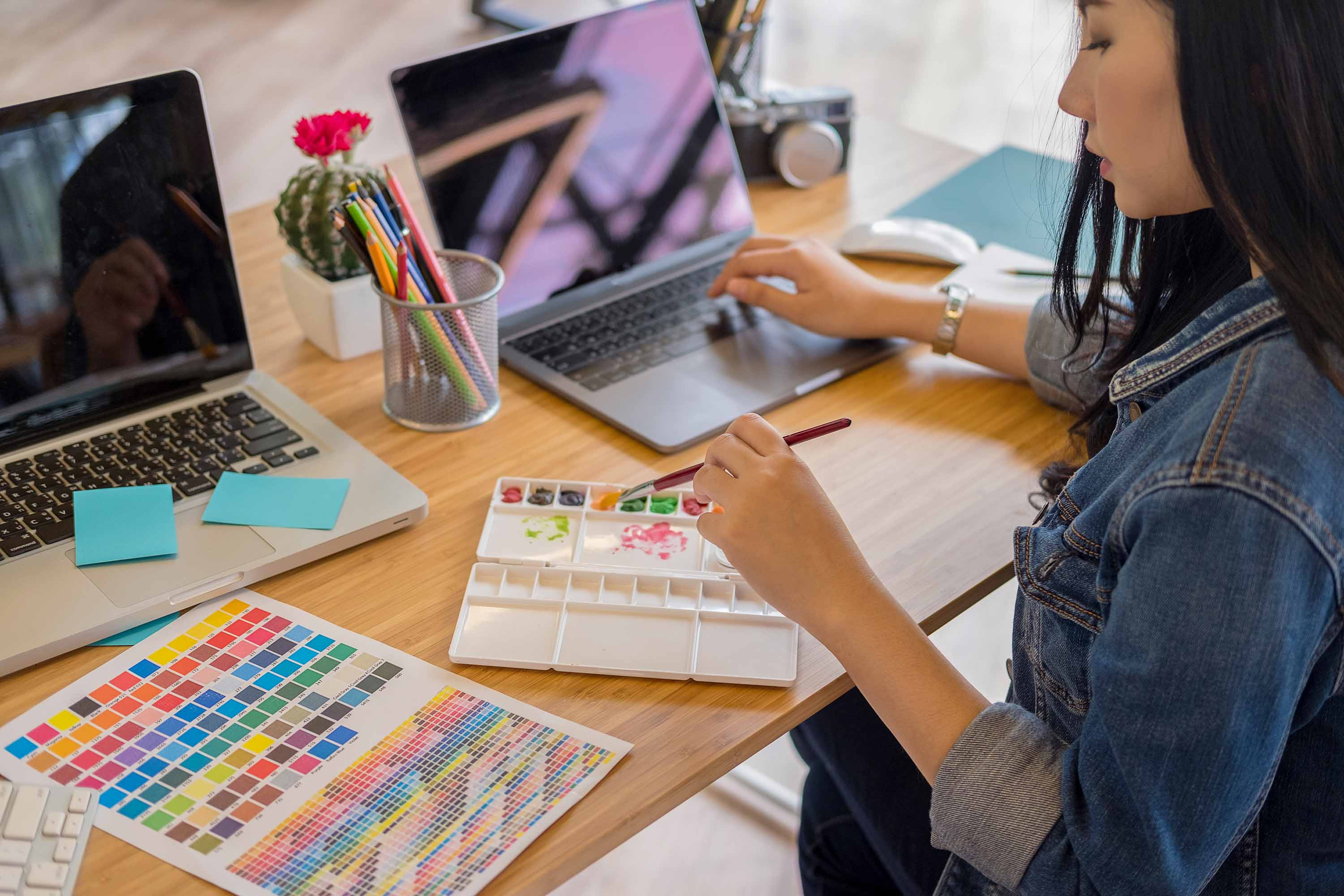 A person mixing watercolour paints whilst interacting with a laptop