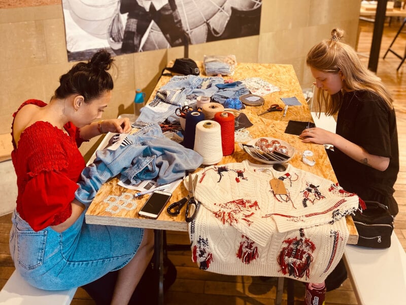 Two women are sitting at a table, mending clothes using the sashiko and boro techniques