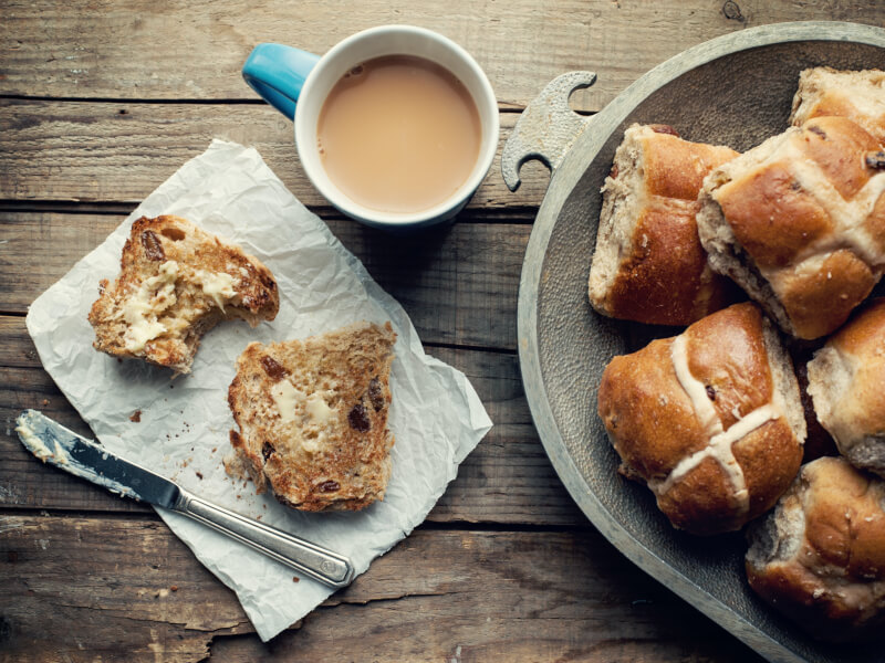 A buttered homemade hot cross bun with a cup of tea next to a plate of more fresh buns. 