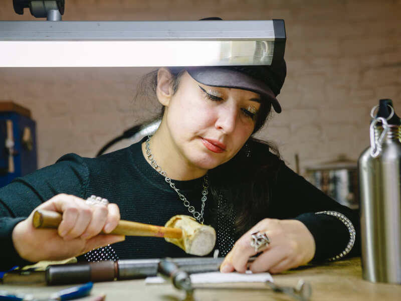 A woman is sitting at a bench, hammering a piece of metal into shape to make a ring