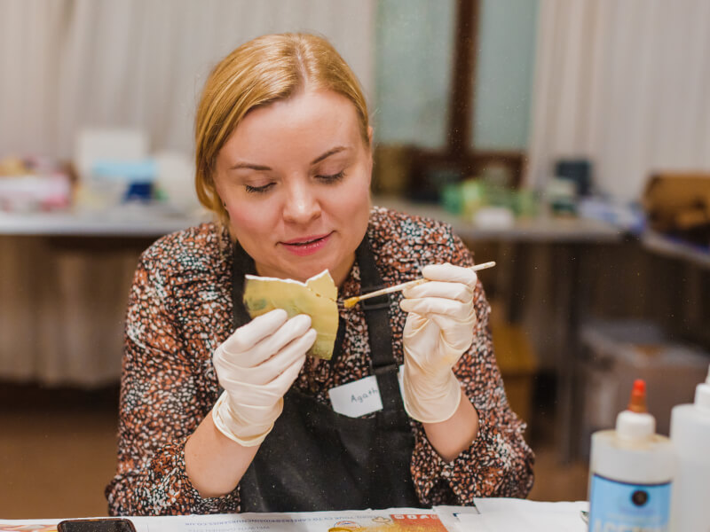 A woman is sitting at a table, applying gold lacquer to a piece of broken plate